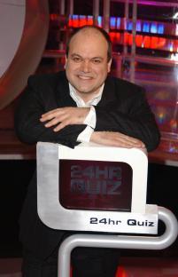A man in a suit leans on a 24hr Quiz set.