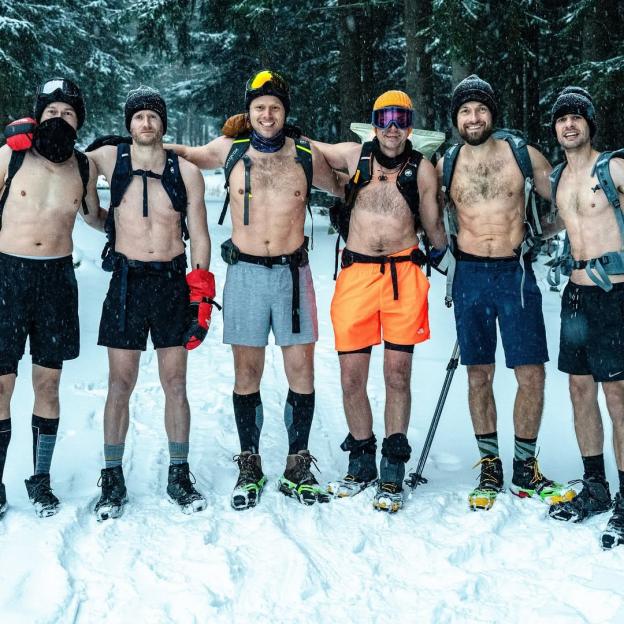 Six men shirtless in shorts and backpacks standing in snow.
