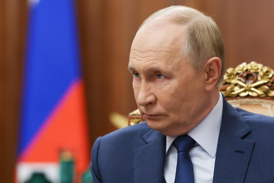 Russian President Vladimir Putin in a suit and tie, looking to his left, with a blurred Russian flag in the background.