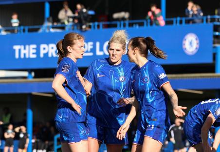 Three Chelsea Women's football players in blue jerseys talking on the pitch.