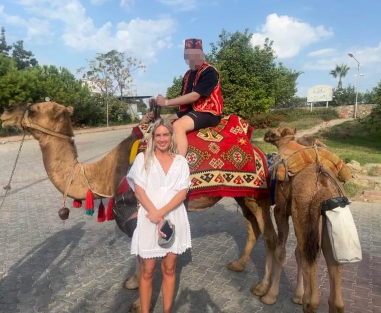 Woman standing next to a camel with a person riding it.
