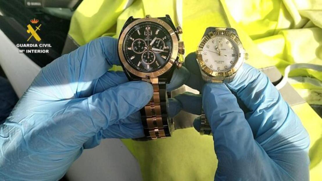 An individual wearing blue gloves holding two watches, one gold-colored with a dark face and one silver with a light face.