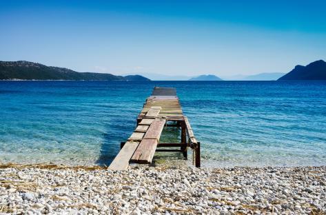 Wooden pier extending into a blue sea, with a pebble beach and mountains in the background.