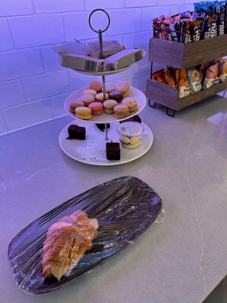 A three-tier stand with snacks, including macarons, brownies, and a dessert cup, with packaged treats in a wooden caddy next to it, and a plastic-wrapped croissant on a plate in the foreground.