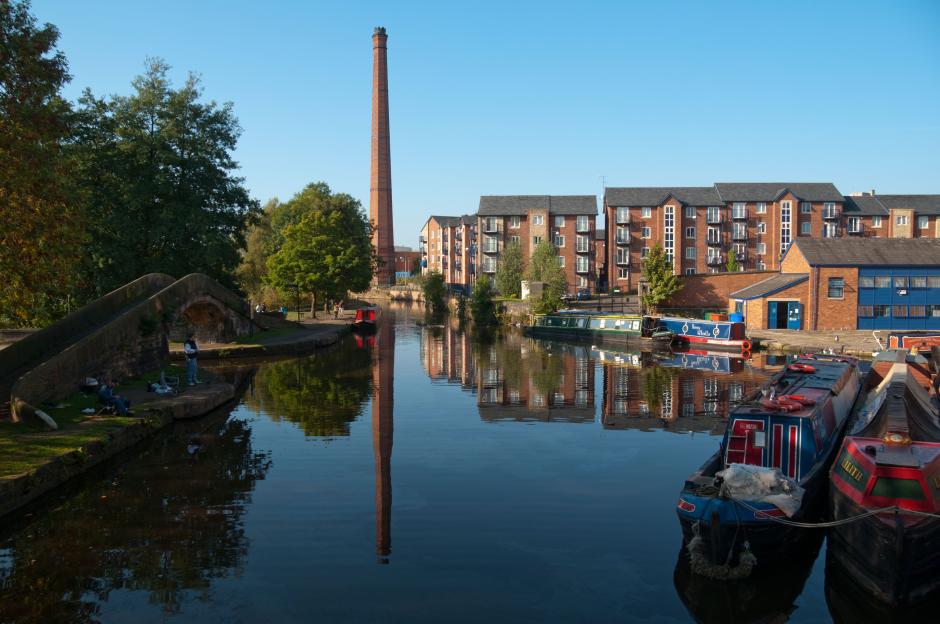 Canal scene with boats, brick buildings, and a tall chimney.