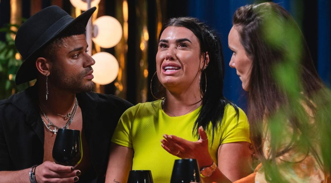 A crying woman in a yellow shirt looks at a man in a black hat, while another woman looks on.