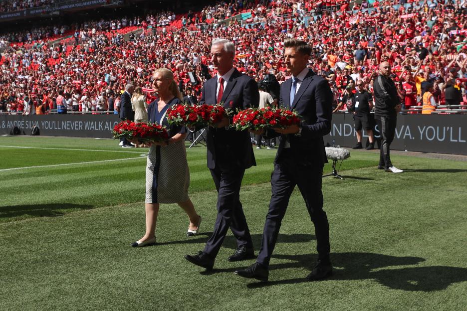 Three people carrying wreaths across a soccer field in front of a stadium crowd.