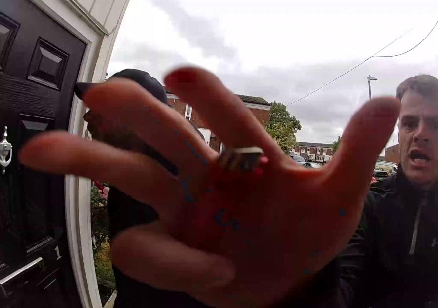 Footage from a doorbell camera showing a man's outstretched hand with a red ring on one finger blocking the camera, and another man with his mouth open to the right.