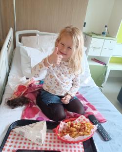 Girl in hospital bed giving thumbs up, eating fries.