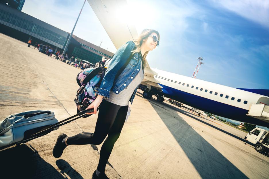 Young woman with suitcase and backpack running on an airport runway next to an airplane.