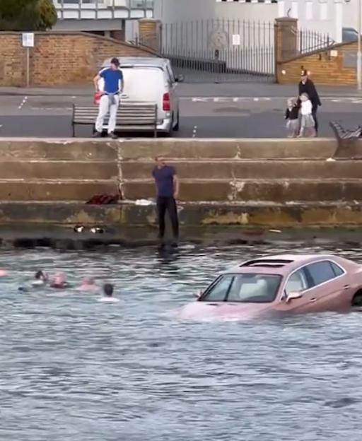 A car partially submerged in water with people nearby in the water and on shore.