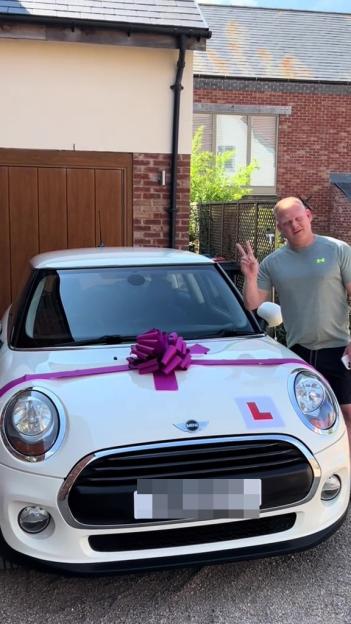 A man stands beside a white Mini Cooper with a purple bow, displaying a learner's permit.