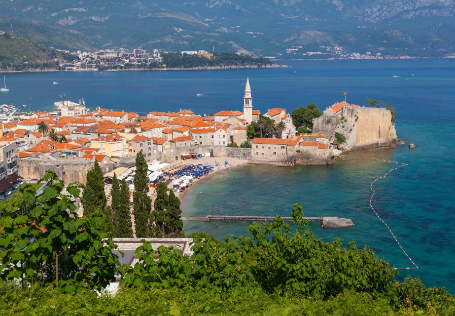 Aerial view of Budva Riviera Resort, featuring a town with red-tiled roofs, a beach, and a clear blue sea.