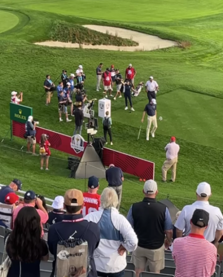 Spectators watch from stadium seats as golfers play on a green course with a sand bunker, surrounded by camera crews.
