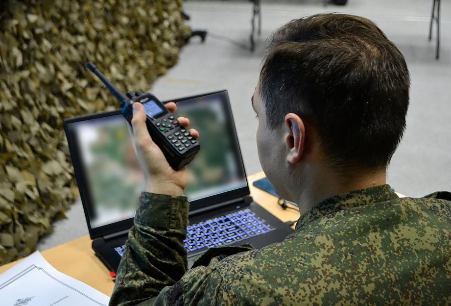 A man in military fatigues holding a walkie-talkie while looking at a laptop screen.