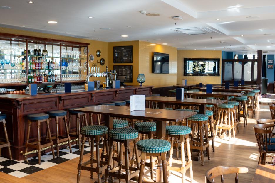 Interior view of The Golden Flake Tavern, a Greggs pub, showing a long bar and multiple high-top tables with stools.