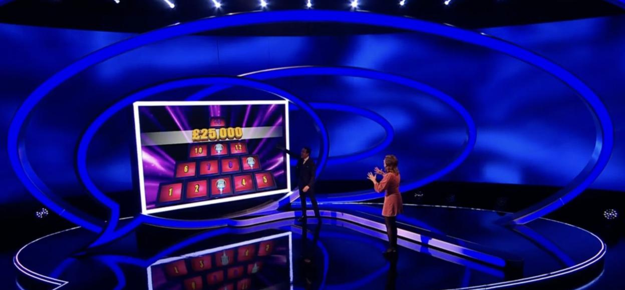Two people on a game show stage, one pointing to a screen with "£25,000" and a grid of numbered panels.
