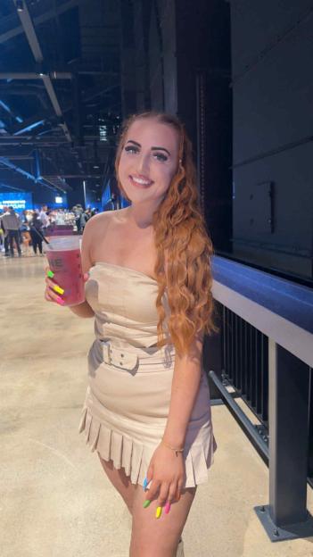 A smiling woman in a beige dress holding a red drink.