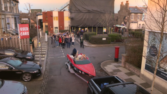 A red speedboat being towed down a street, with people watching.