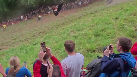 A person back-flipping down a hill at a cheese rolling competition, with spectators recording the event on their phones.