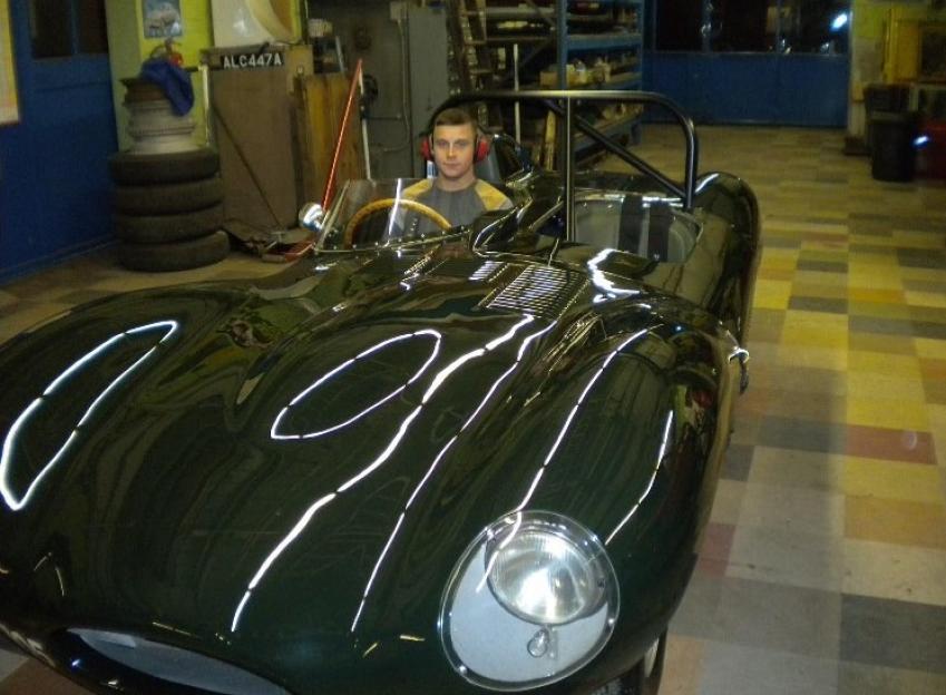 Man sitting in a dark green Jaguar D-Type sports car in a garage.