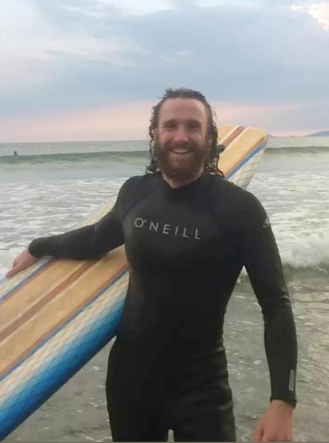 Photo of Alexander Barrett, smiling, wearing a wetsuit and holding a surfboard.