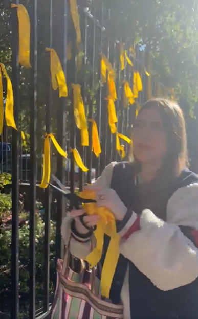 A woman tying a yellow ribbon to a black metal fence, with many other yellow ribbons already attached.