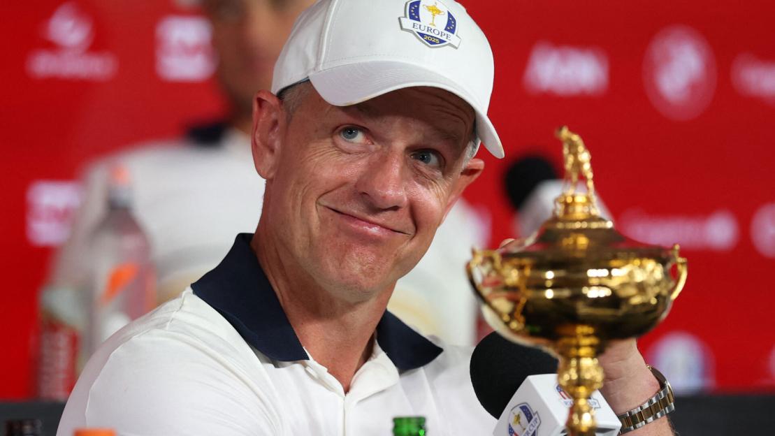 Luke Donald wearing a white "Europe 2025" cap, smiling next to the Ryder Cup trophy.