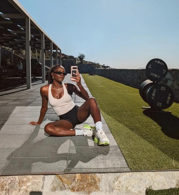 AJ Odudu taking a selfie while sitting on a tiled surface next to a grassy area with weight plates.