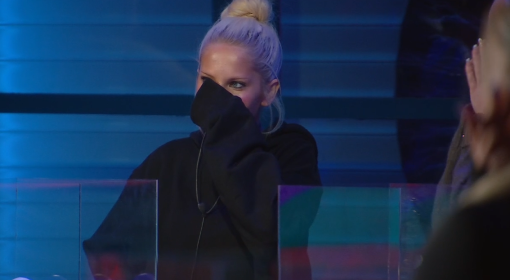 A woman in a black hoodie with blonde hair in a bun covering her mouth with her sleeve, her eyes visible above her arm.