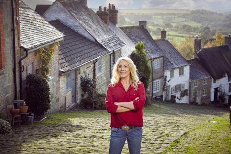 Sarah Beeny standing in a cobblestone street in front of old houses.