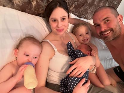 Family of five in bed; parents hold newborn and toddler, while older daughter sits between them.