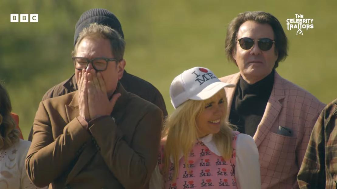 The cast of "The Celebrity Traitors" watching something shocking, with one man covering his mouth and a woman in an "I ♥ Me" shirt smiling.