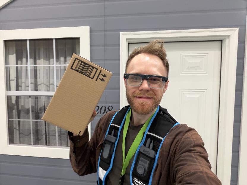 A smiling man wearing smart glasses and an Amazon vest holds up a cardboard box in front of a house facade.
