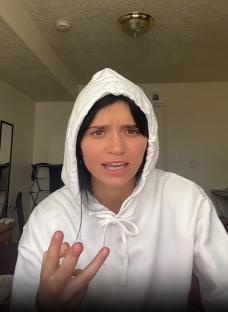 Katie Mills in a white hoodie, making a hand gesture.