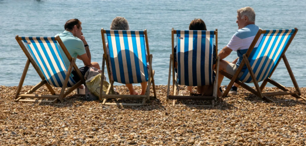 Four people relaxing in deck chairs on a pebble beach, facing the sea.