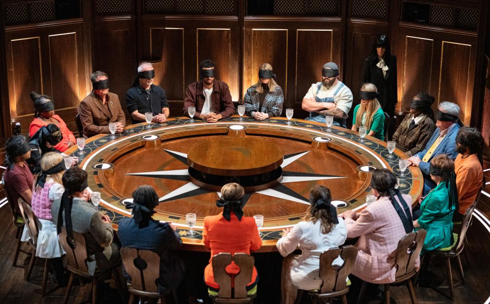 A group of contestants with blindfolds seated around a circular table in a dimly lit room.