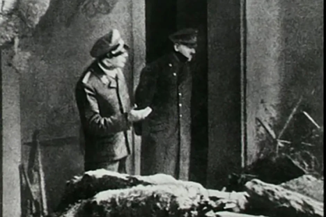 Black and white photo of two men in uniform standing near a pile of bodies.