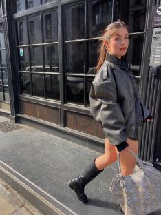 Woman walking down the street wearing a black leather jacket and tall black boots, carrying a light-colored handbag.
