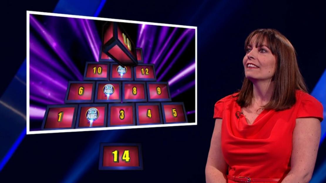 A woman in a red dress smiles next to a screen showing a pyramid of numbered boxes from the game show Catchphrase.