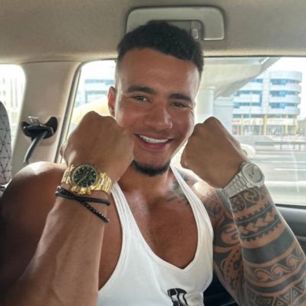 George Fensom smiling with fists raised, showcasing two watches and arm tattoos.