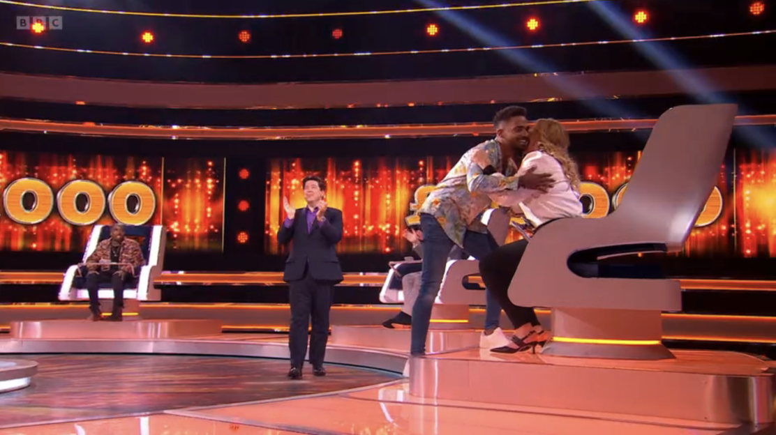 Screenshot of a game show; contestants hugging while a host applauds.
