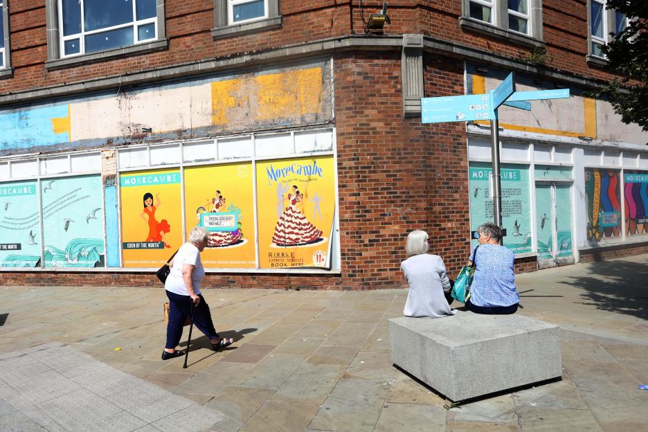 Two women sit on a low stone bench, watching an older woman walk with a cane past boarded-up shops with colorful posters advertising Morecambe.