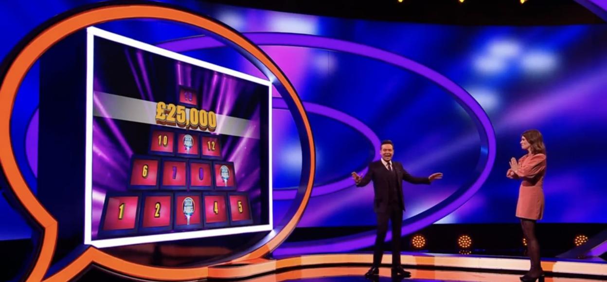 A game show host and comedian Ellie Taylor standing on a brightly lit stage in front of a game board showing £25,000.