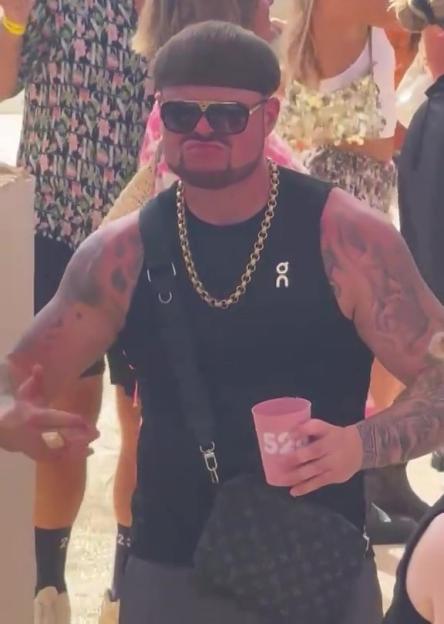 A man with a bowl cut, sunglasses, gold chain, and tattoos holds a pink cup at a party.