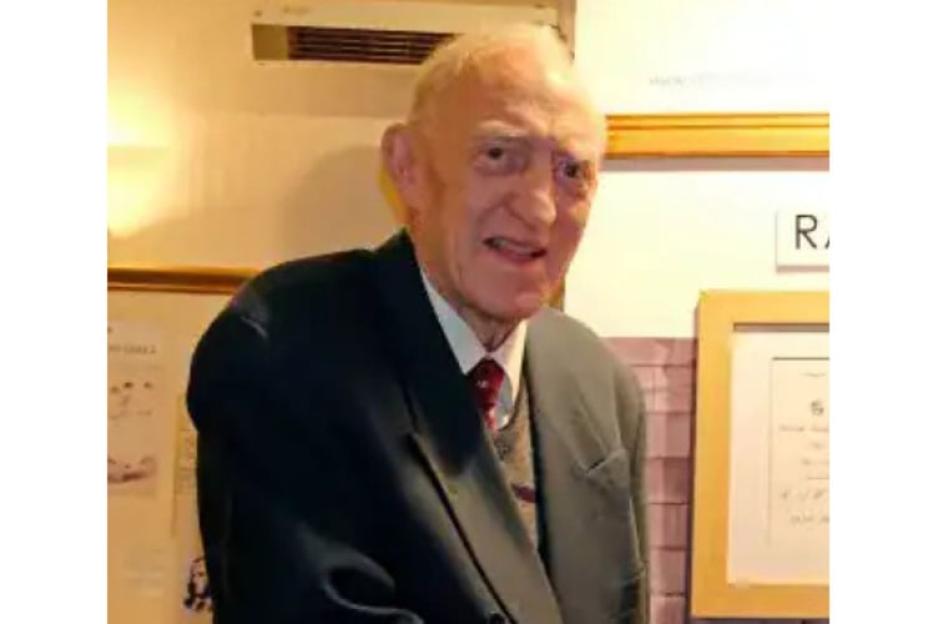 A man in a suit jacket and tie looks at the camera, with framed documents behind him.