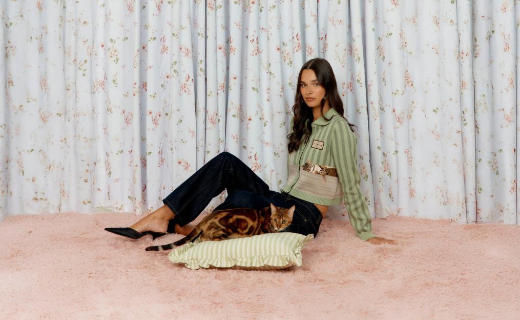 A woman sitting on a pink rug with a Bengal cat, wearing a green striped cropped cardigan.
