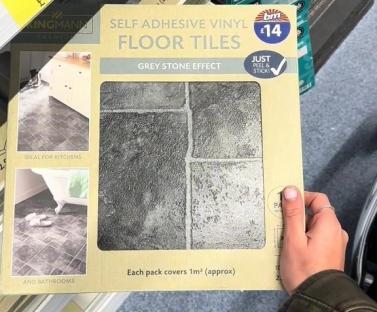 Person holding a box of grey stone effect self-adhesive vinyl floor tiles.