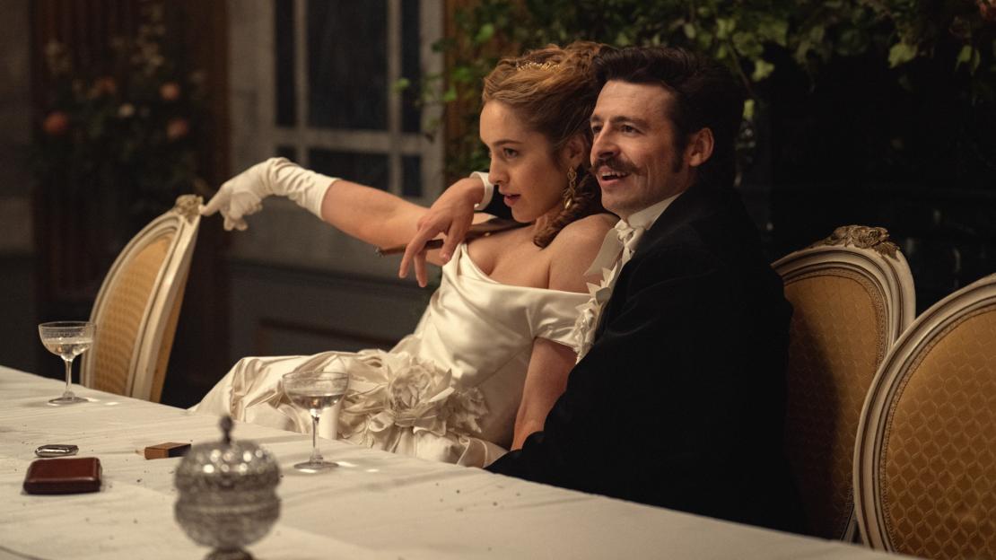 A man and a woman in formal attire at a table, with the woman holding a cigar.