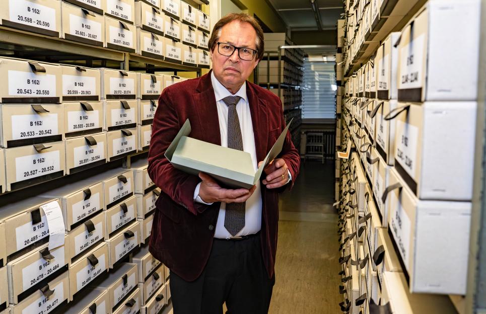 Senior Public Prosecutor Thomas Will standing in an archive with rows of labeled boxes and holding an open file.